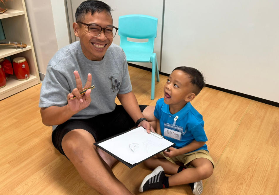 Smiling man flashes peace sign while holding pencil; young boy beside him holds a drawing board on the floor.